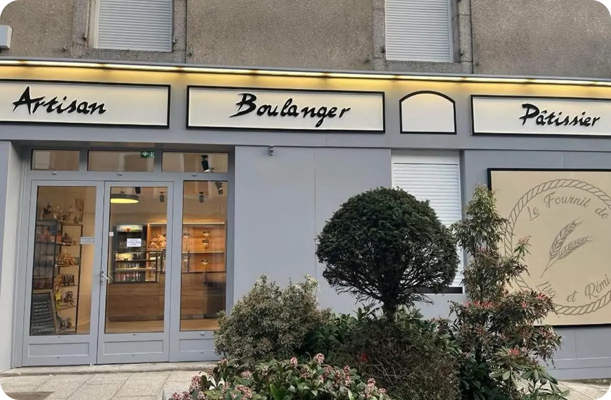 La devanture d'une boulangerie française avec des panneaux indiquant Artisan Boulanger Pâtissier. L'entrée a des portes vitrées et la fenêtre montre des étagères avec des produits de boulangerie. Des buissons et des fleurs se trouvent devant la boutique.