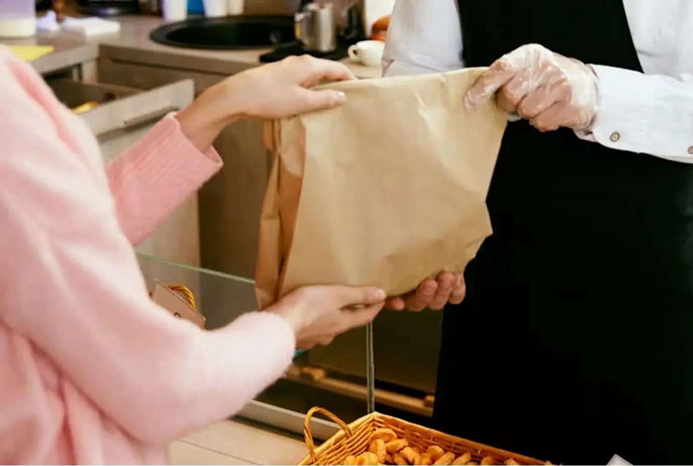 Une cliente reçoit un sac en papier kraft d’un vendeur en tablier noir et gants jetables, au comptoir d'une boulangerie. Un panier garni de petits pains est visible au premier plan.