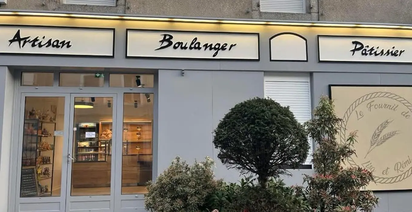 Une charmante devanture de boulangerie française avec des panneaux indiquant Artisan Boulanger Pâtissier à Brassac. De grandes vitrines présentent les produits de boulangerie et un arbre et des arbustes vous accueillent à l'entrée.