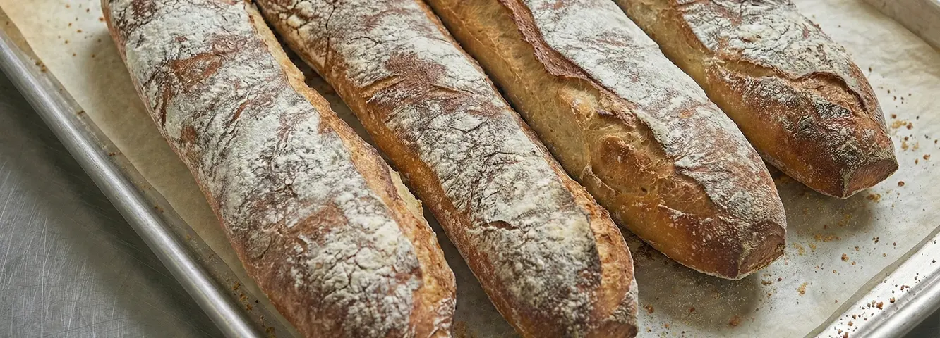 Pain rustique fraîchement cuit, disposé sur une plaque de cuisson. Les baguettes présentent une croûte dorée et farinée, avec des grignes bien ouvertes.