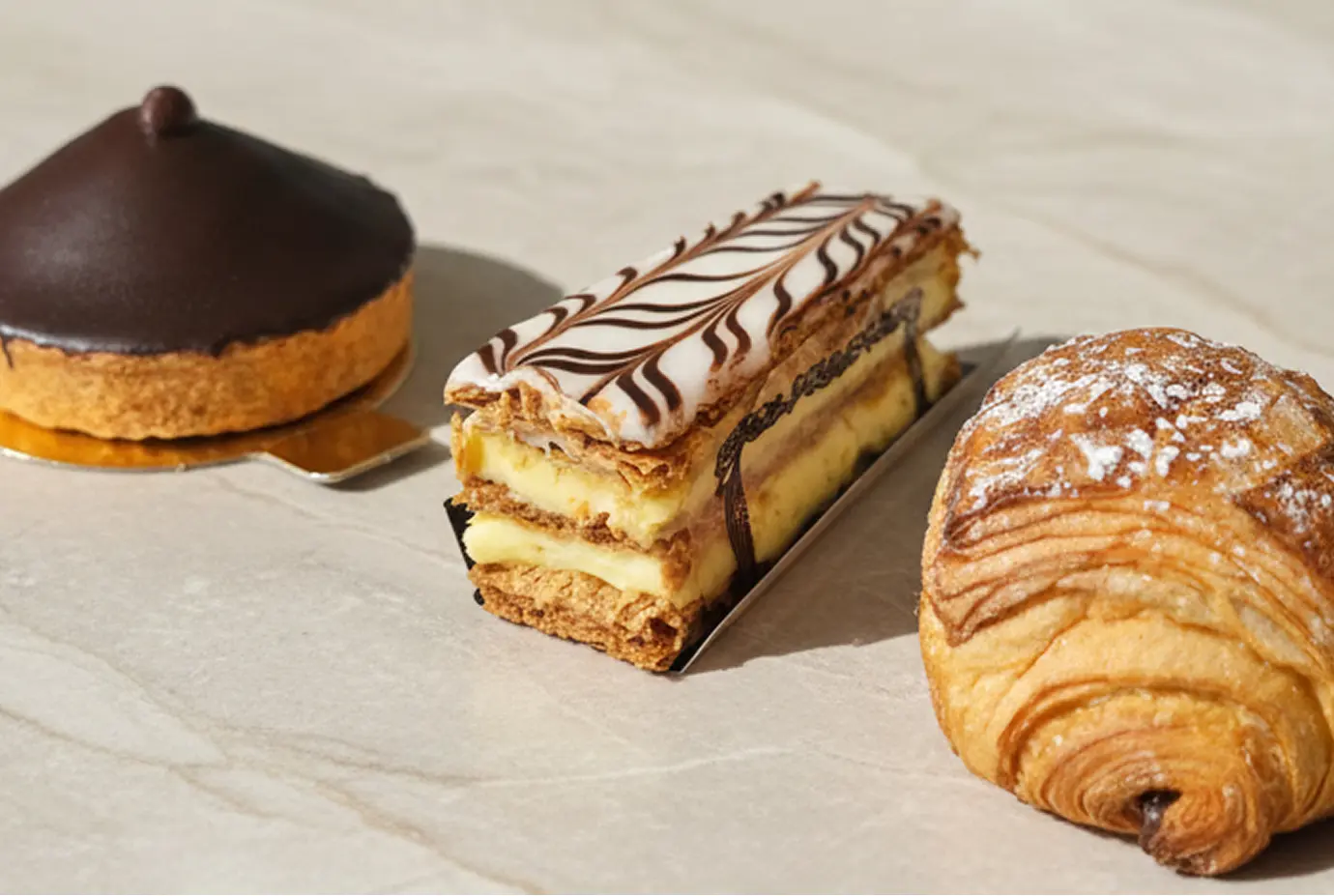 Assortiment de pâtisseries françaises : une tartelette chocolatée, un mille-feuille à la crème vanille et un pain au chocolat saupoudré de sucre glace, présentés sur un plan en marbre clair.