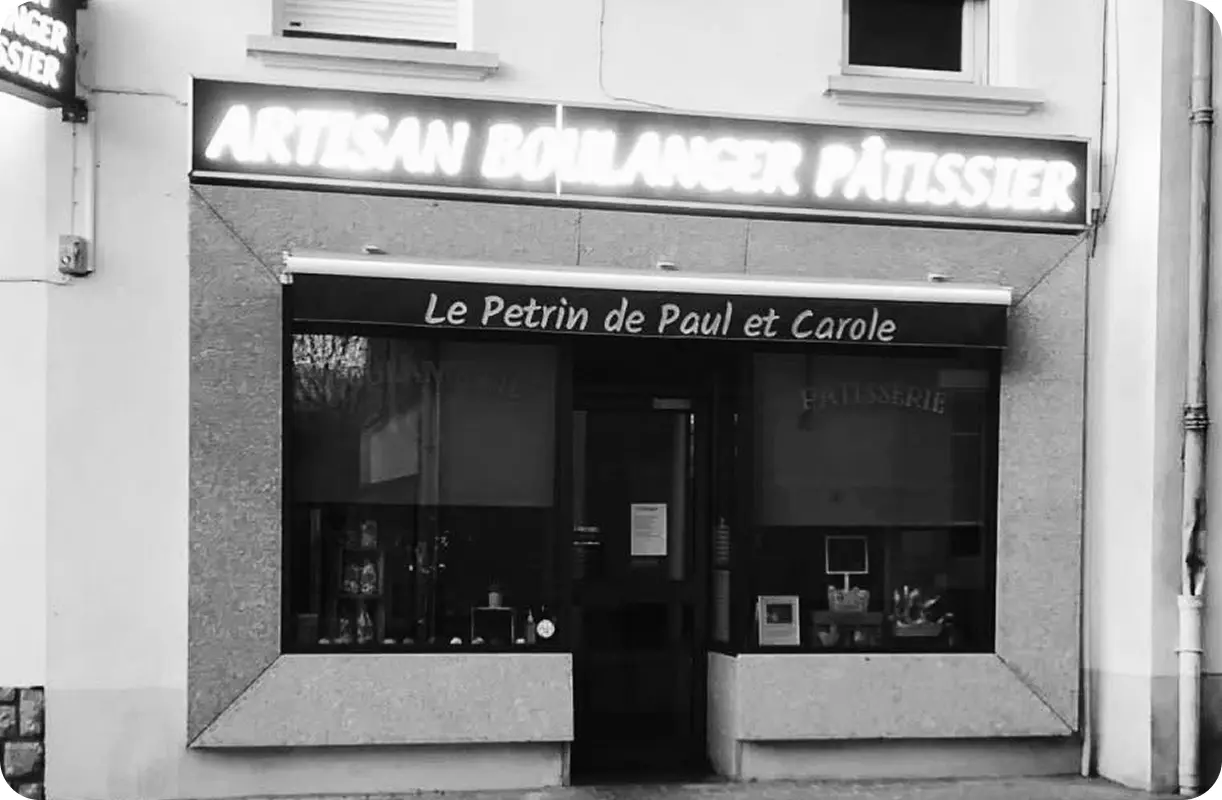 Photo en noir et blanc de la devanture d'une boulangerie avec une enseigne lumineuse en néon indiquant "ARTISAN BOULANGER PÂTISSIER". La vitrine en dessous affiche "Le Petrin de Paul et Carole". Des articles sont visibles dans la vitrine.