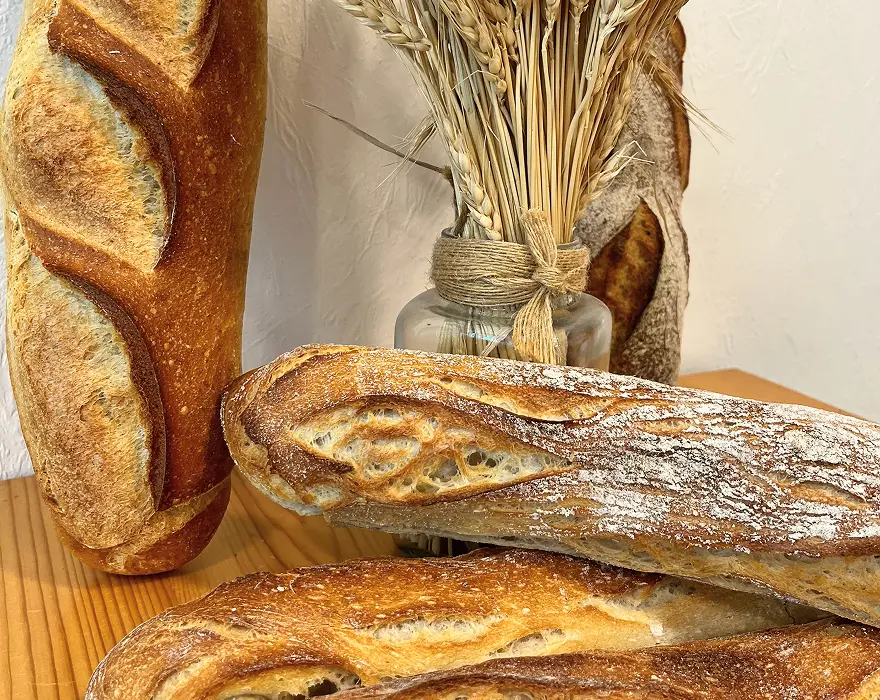 Assortiment de pains artisanaux à la croûte dorée et aux grignes marquées, posés sur une surface en bois, avec un bouquet d’épis de blé dans un bocal en verre décoré de ficelle, en arrière-plan.