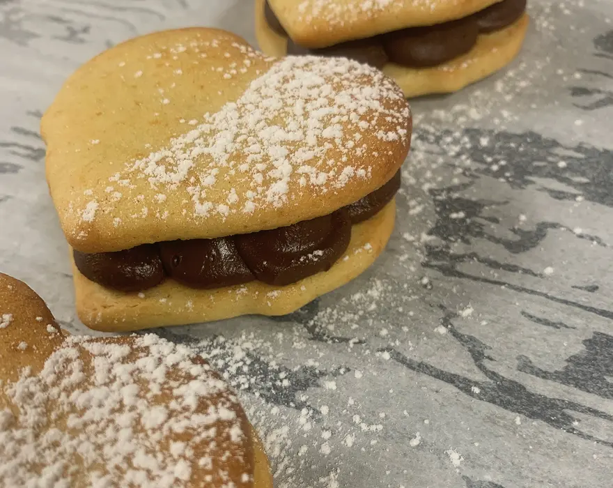 Biscuits sablés en forme de cœur, garnis d’une ganache chocolatée entre deux couches, et saupoudrés de sucre glace sur le dessus, disposés sur une feuille de cuisson.