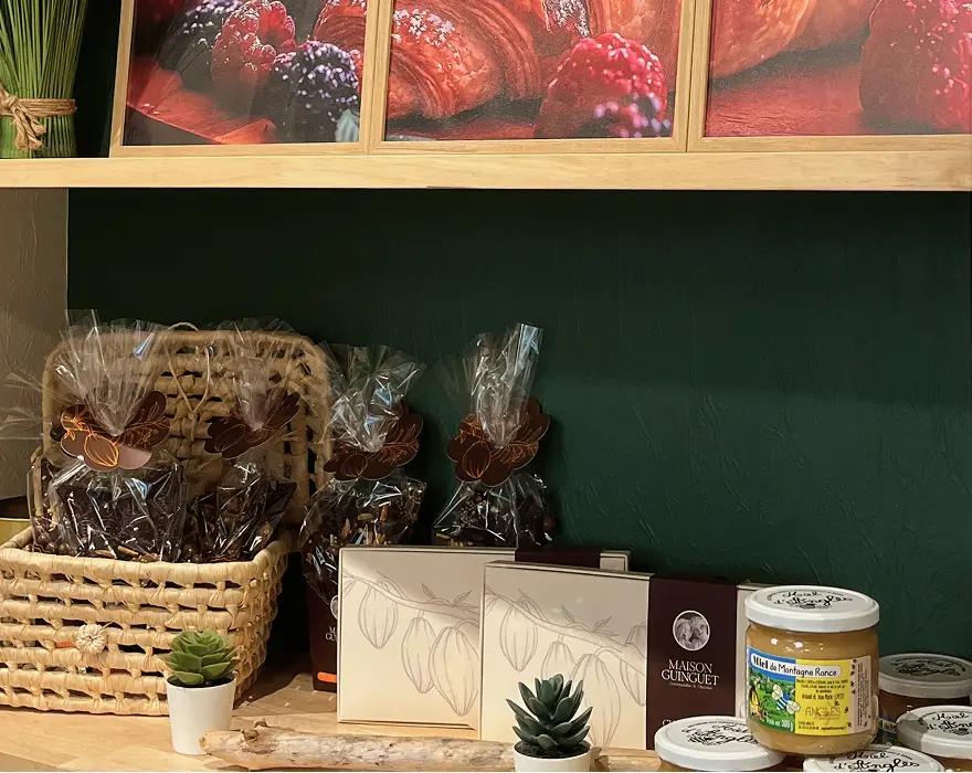 Étagère de boutique garnie de sachets de chocolats emballés, de boîtes élégantes avec dessins de cabosses de cacao, de pots de miel, de petits pots décorés, et de plantes miniatures, avec en fond une photo de pâtisseries aux fruits rouges.