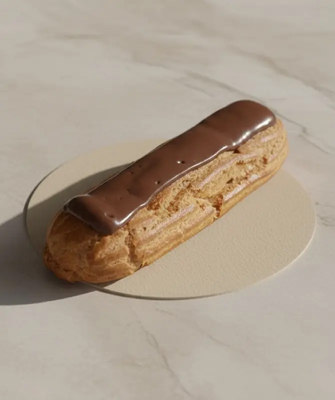 Éclair au chocolat posé sur un support rond beige, avec une pâte à choux dorée et un glaçage brillant au chocolat, éclairé par une lumière naturelle sur fond en marbre clair.