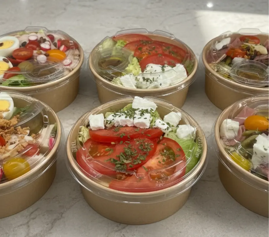 Assortiment de salades composées individuelles dans des bols en carton avec couvercles transparents, garnies de légumes frais, tomates, fromage, œufs, olives, et sauce séparée, présentées sur un plan de travail clair.