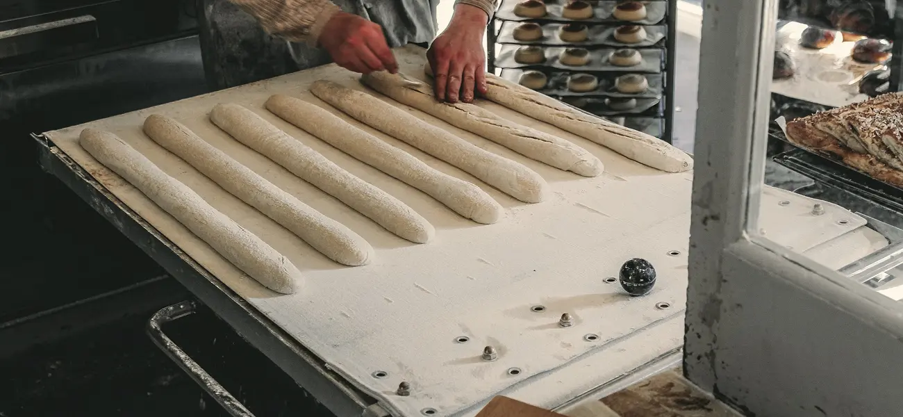 Un boulanger façonne des baguettes crues sur un tapis de fermentation fariné, en incisant la pâte à la lame avant la cuisson. À l’arrière-plan, des pains et viennoiseries attendent sur des grilles.