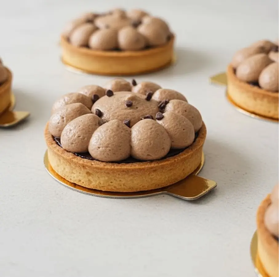 Tartelettes individuelles garnies de crème mousseuse au chocolat, dressée en grosses gouttes sur une base de pâte sablée dorée, avec quelques pépites de chocolat en décoration, disposées sur un fond clair.