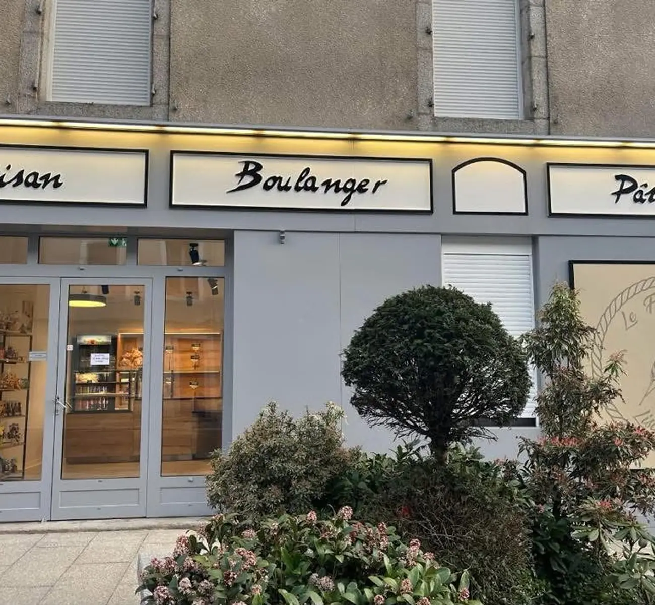 Une charmante devanture de boulangerie française avec des panneaux indiquant Artisan Boulanger Pâtissier à Brassac. De grandes vitrines présentent les produits de boulangerie et un arbre et des arbustes vous accueillent à l'entrée.