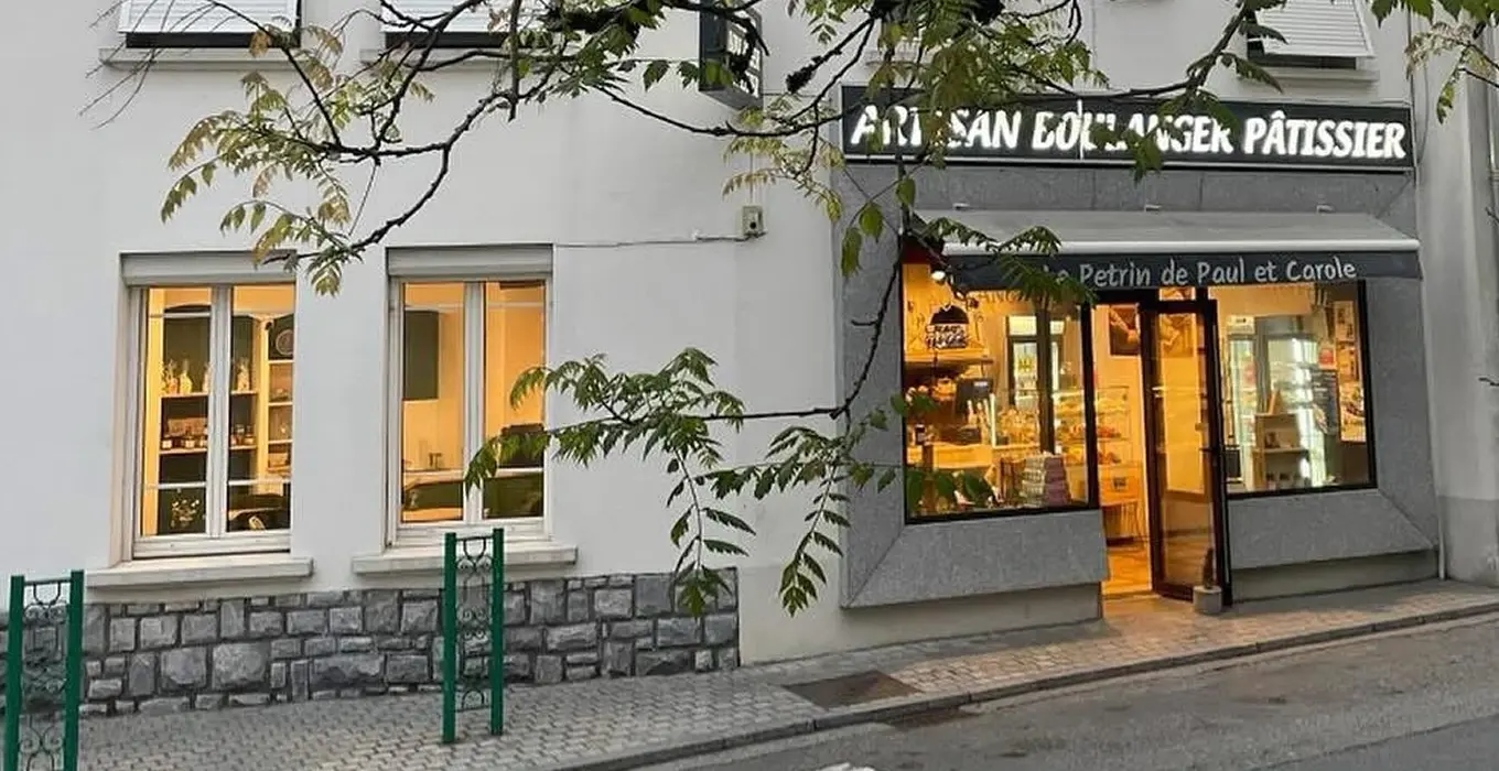 Façade d’une boulangerie-pâtisserie artisanale nommée Le Pétrin de Paul et Carole, avec vitrine éclairée et enseigne lumineuse “Artisan Boulanger Pâtissier”, située dans une rue calme avec passage piéton et arbres au premier plan.