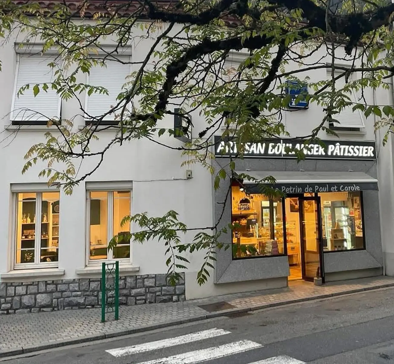 Façade d’une boulangerie-pâtisserie artisanale nommée Le Pétrin de Paul et Carole, avec vitrine éclairée et enseigne lumineuse “Artisan Boulanger Pâtissier”, située dans une rue calme avec passage piéton et arbres au premier plan.