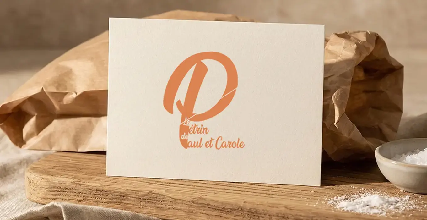 Carte posée sur une planche en bois avec le logo « Le Pétrin de Paul et Carole » imprimé en orange, devant un sac en papier kraft froissé et un petit bol de farine, sur fond de tissu beige et décor naturel.