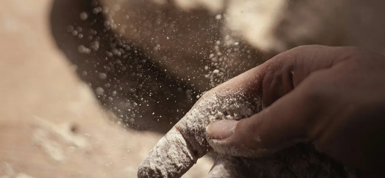 Main d’un boulanger saupoudrée de farine en plein geste, avec des particules de farine en suspension dans l’air, éclairées par une lumière douce, évoquant la précision et l’artisanat du travail du pain.